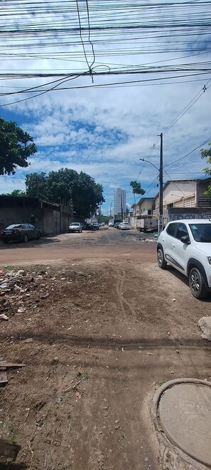 Uma rua com um carro branco estacionado à direita, edifícios ao fundo e uma área de terra à frente.