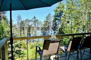 Eine Holzterrasse mit Stühlen und einem Sonnenschirm, mit Blick auf einen Wald und ein Gewässer.
