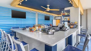 Una zona de bar con una barra alargada, taburetes, ventiladores de techo y estantes con botellas.
