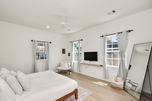 Un dormitorio con cama, una silla, televisión, espejo y ventanas con cortinas.