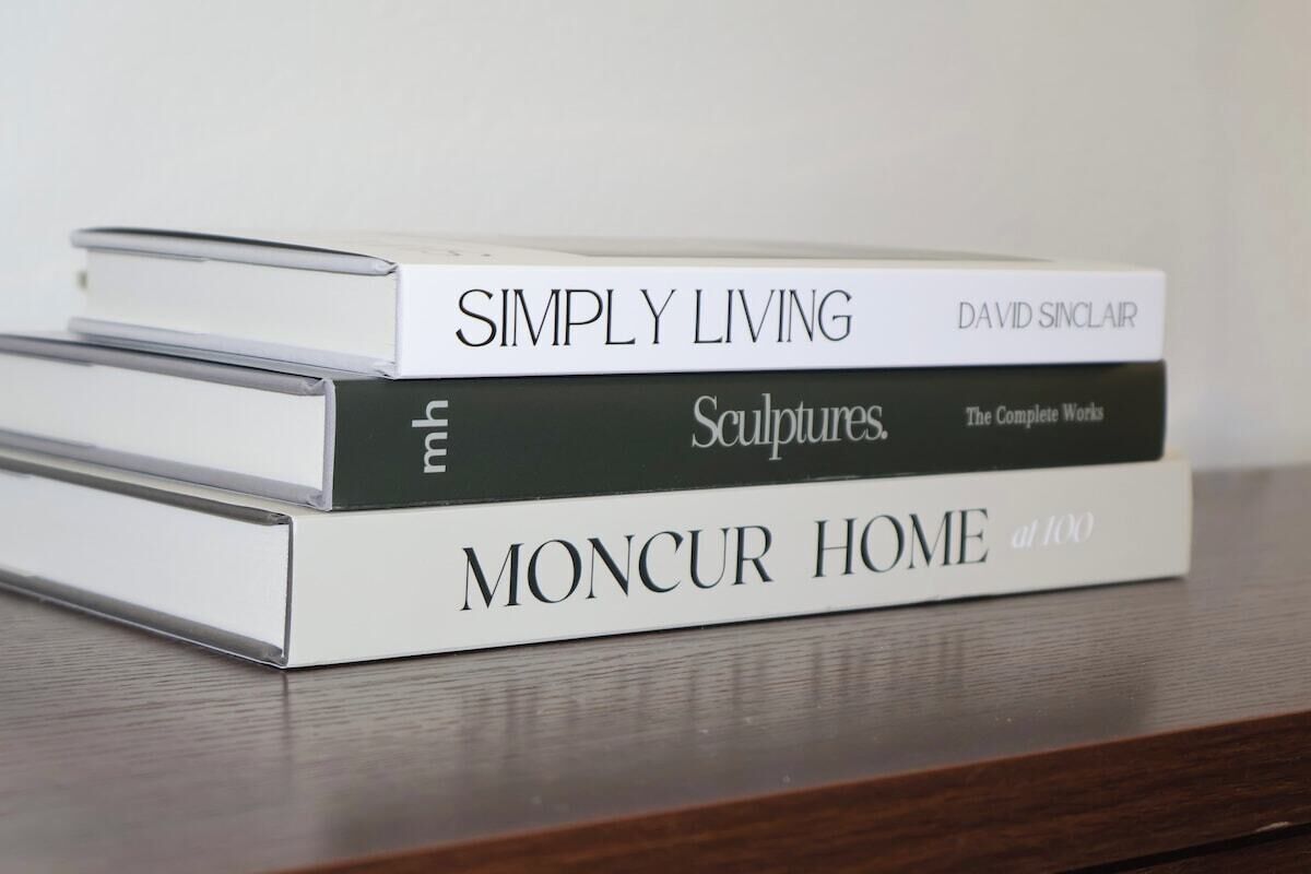 A stack of books titled 'Simply Living' by David Sinclair, 'Sculptures' by mh, and 'Moncur Home at 100' on a wooden surface.