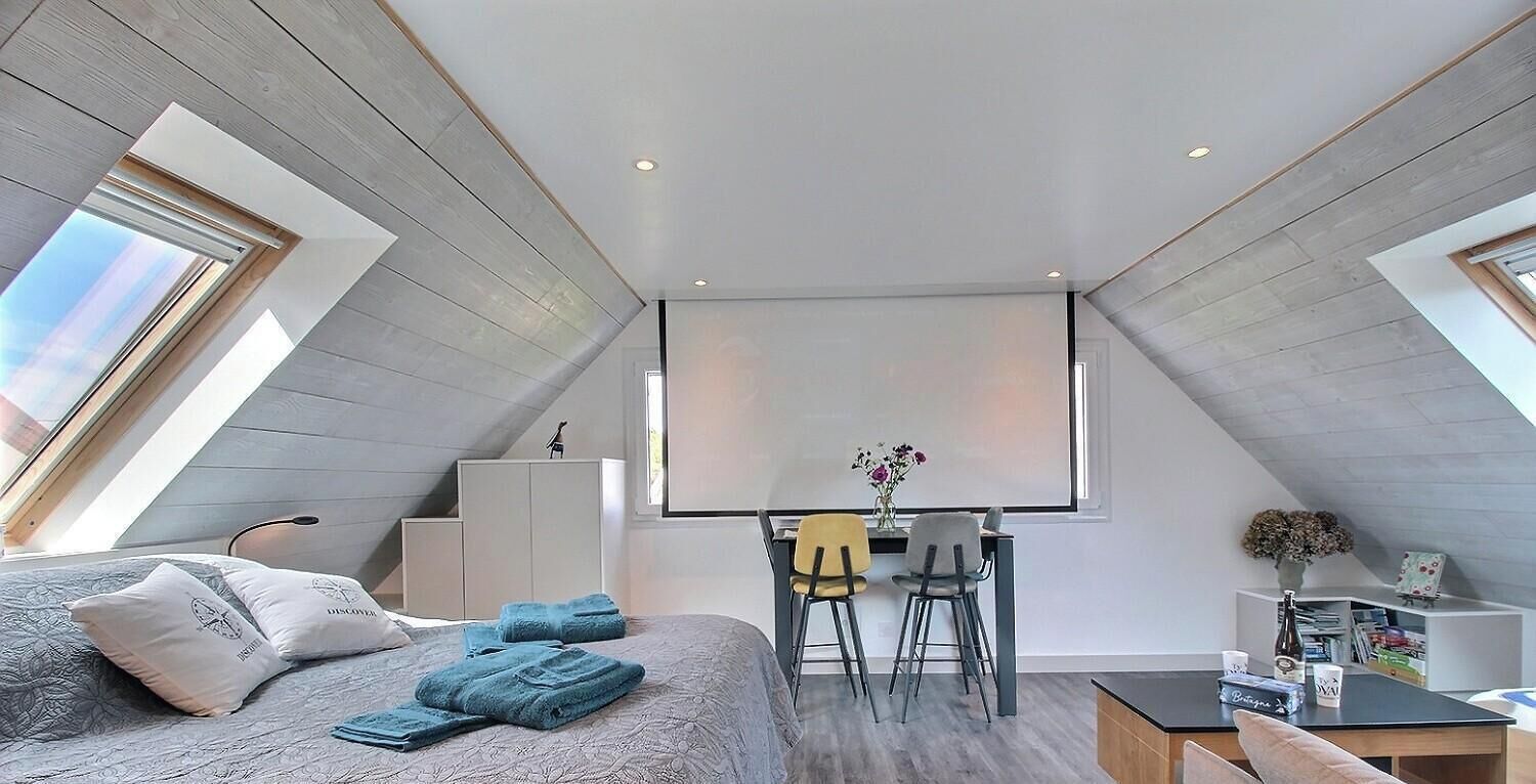 Un dormitorio estilo loft moderno con tragaluces, una cama con ropa de cama estampada, un escritorio con dos sillas y una mesita con un jarrón.