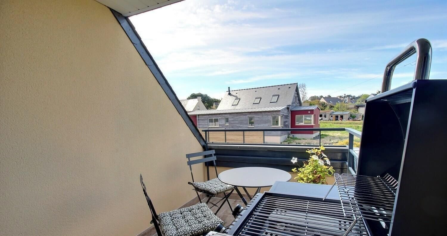 A balcony with a table and chairs, a grill, and a view of houses in the background.