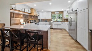 A modern kitchen with white cabinets, a large island with bar stools, and a view of greenery outside. - Sunrise Mountain Views, Hot Tub, Fire Pit & Spacious Retreat Near Downtown Boone (Boone)