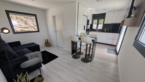 Een moderne woonkamer met een bank, een eethoek met krukken, een keuken met witte kastjes en een groot raam met uitzicht op een rotsachtig landschap.