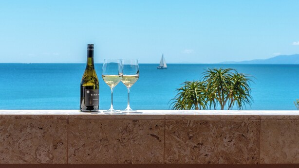 A bottle of wine and two glasses on a ledge with a view of the sea and a sailboat. - Vista On The Beach at The Sands (Waiheke Island)