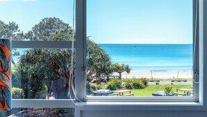 A view from a window overlooking a beach with a clear sky, a grassy area, and a few trees. - Vista On The Beach at The Sands (Waiheke Island)