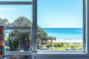 A view from a window overlooking a beach with a clear sky, a grassy area, and a few trees. - Vista On The Beach at The Sands (Waiheke Island)
