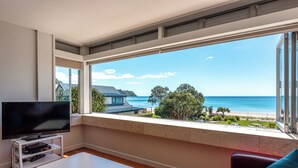A room with a large window, a flat-screen TV on a white stand, a white table, and a red couch with a blue pillow. - Vista On The Beach at The Sands (Waiheke Island)