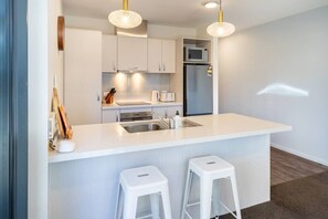 Eine moderne Küche mit weißen Schränken, einer Edelstahlspüle, einer Mikrowelle und einem Kühlschrank. Zwei weiße Barhocker und eine kleine Kücheninsel mit Spüle gehören ebenfalls dazu.