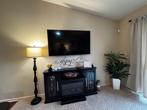 A living room with a flat-screen TV mounted on the wall, a lamp, a console table with decorative items, and a potted plant.