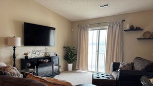 A living room with a flat-screen TV, a fireplace, a large window with curtains, and a potted plant. - 2-bedroom 2-bath condo in lovely Mishawaka close to Notre Dame. (Mishawaka)