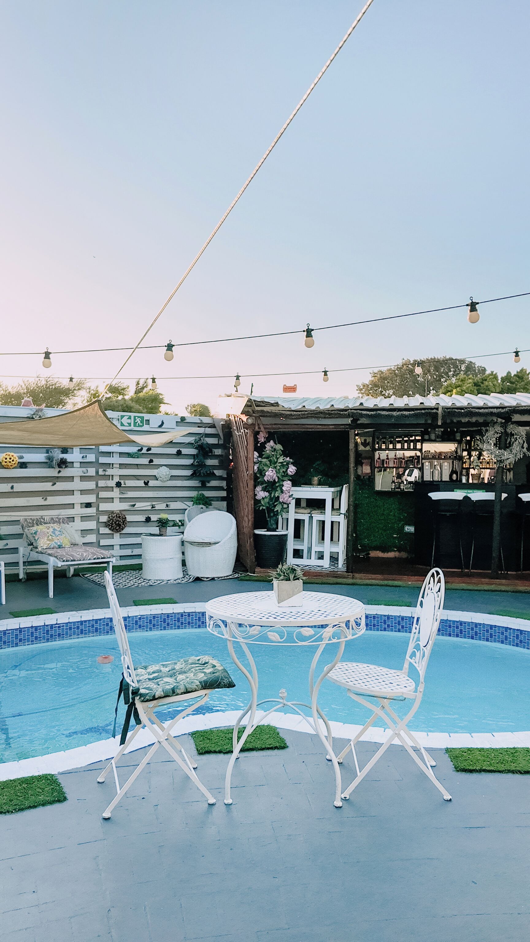 Un espace piscine avec des chaises et une table en métal blanc, entouré d’une clôture agrémentée de plantes, et un bâtiment visible au fond.