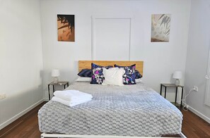 Una cama bien hecha con un edredón estampado, dos mesitas de noche con lámparas y dos obras de arte colgadas en la pared.
