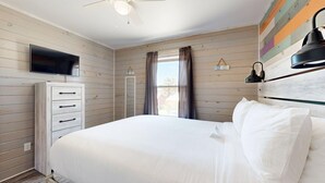 Une chambre à coucher avec un lit, une commode, un téléviseur, un ventilateur de plafond et une fenêtre avec des rideaux.