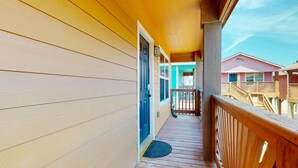 Une terrasse en bois avec une porte bleue, une balustrade en bois et, en arrière-plan, une vue sur des bâtiments aux couleurs vives.
