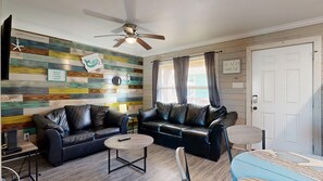 Ein Wohnzimmer mit einer Wand aus Holzplanken, einem Deckenventilator, einer schwarzen Ledersofa, einem runden Couchtisch und einer Tür mit der Aufschrift 'BEACH HOUSE'.
