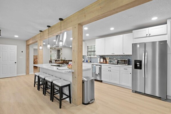 A modern kitchen with white cabinets, stainless steel appliances, a central island with bar stools, and wooden beams. - Sleeps Ten, Pet friendly, White sand beaches (Pensacola)