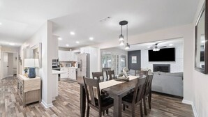 Una cocina moderna con armarios blancos, un comedor con mesa y sillas de madera, y una sala de estar con sofá gris y chimenea.