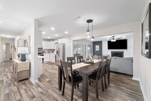 A modern kitchen with white cabinets, a dining area with a wooden table and chairs, and a living area with a grey sofa and fireplace. - Sea`s the Day Getaway (Pensacola)