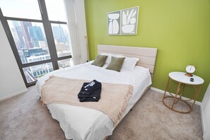Un dormitorio con una cama, una mesa redonda y vistas a un paisaje urbano a través de amplios ventanales.