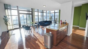 Une cuisine moderne dotée d’un grand îlot central, d’un coin repas avec des chaises et offrant une vue sur la ville grâce à de grandes fenêtres.
