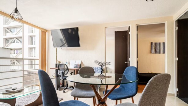 Un salón moderno con una mesa de vidrio, sillas azules y grises, un televisor de pantalla plana y vistas de un edificio a través de amplias ventanas.