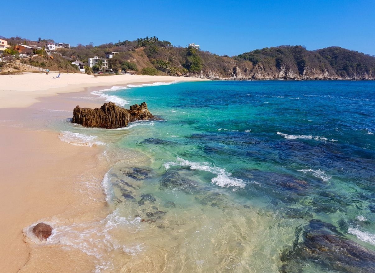 A beach with clear turquoise water, a rocky outcrop, and a hillside with buildings.