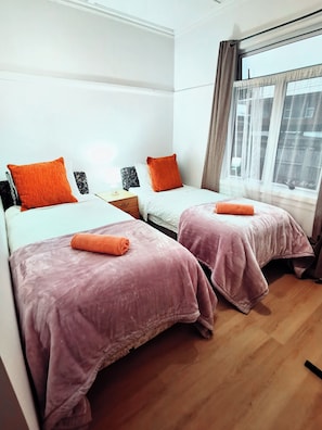 Een kamer met twee bedden, elk met een roze sprei en een oranje kussen. Daartussen staat een houten nachtkastje met een lamp.