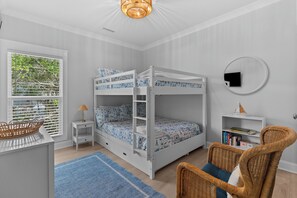A child's bedroom with bunk beds, a small side table, a wicker chair, a bookshelf, and a window with blinds. - Saltwater Pool/Putting Green + Minutes to Beach (Oak Island)