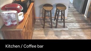 Une cuisine avec un coin café, des étagères en bois, un réfrigérateur et une pancarte sur laquelle on peut lire : « Que cette maison soit bénie. »