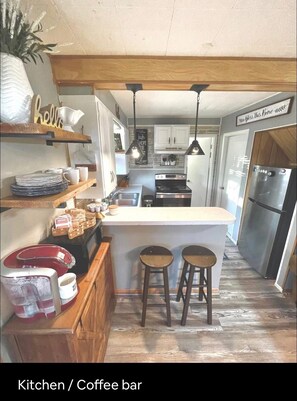 Une cuisine avec un coin café, des étagères en bois et un panneau sur lequel on peut lire : « Que cette maison soit bénie. »