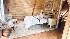 Une chambre avec un plafond en pente, des murs en bois, un lit avec une literie blanche, une table de chevet, une chaise et un luminaire fixé au mur.