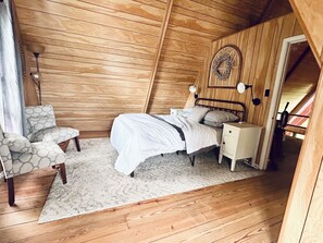 A bedroom with a wooden sloped ceiling, a bed with white bedding, a nightstand, a chair, and a wall-mounted light fixture.
