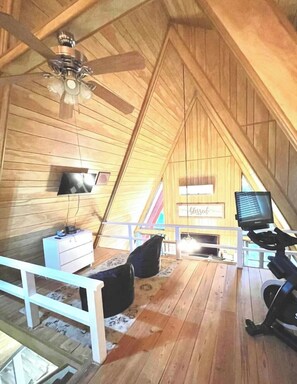 Une pièce avec un plafond en pente, des murs en bois, un ventilateur de plafond, une télévision, un bureau et un ordinateur.