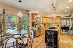 A modern kitchen with wooden cabinets, a dining area with a round table and chairs, and a view of the outdoors through large windows. - Fossil Run! Curated Home w/ Wellness & Game Room (Colorado Springs)