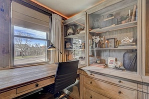 A room with a desk, chair, and a large window. There is a bookshelf with various items on it.