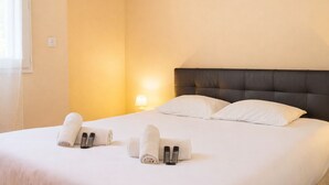 A hotel room with a bed, pillows, a nightstand with a lamp, and rolled towels. - L’Oasis Helvétie - Faron View - Welkeys (Toulon)