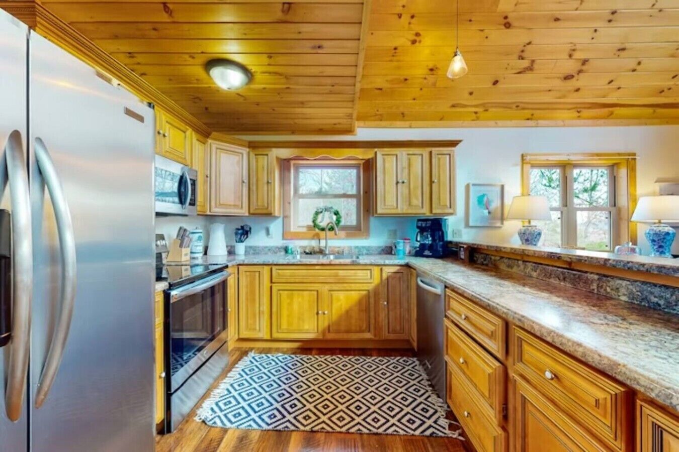 A kitchen with wooden cabinets, a stainless steel refrigerator, a patterned rug, and a window with a wreath.