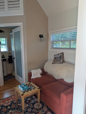 Ein Wohnzimmer mit einem roten Sofa, einem kleinen Tisch, einem gemusterten Teppich, einem Fenster mit Jalousien und einer Wandleuchte.