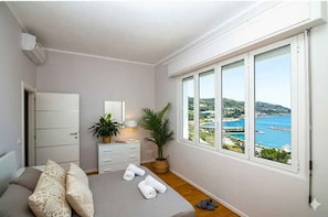 Ein Schlafzimmer mit einem Bett, einer Kommode, einem Nachttisch, einer Topfpflanze und Blick auf eine Küstenlandschaft durch große Fenster.