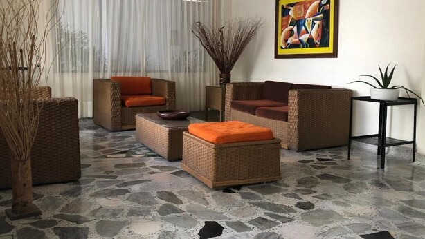 A living room with wicker furniture, a stone floor, a large window with sheer curtains, and a framed abstract painting. - Hotel Mar Azul by Croma (Santa Marta)