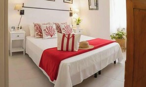 Ein ordentlich bezogenes Bett mit einer roten und weißen Tagesdecke, weiße Kissen mit roten Baum-Motiven und ein kleiner runder Tisch mit einer Lampe und einer Pflanze.