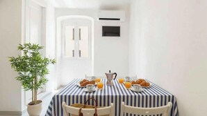 Ein Essbereich mit gestreiftem Tischläufer, zwei Stühlen, einer Zimmerpflanze und Frühstücksangeboten wie Croissants, Orangensaft und Kaffee.
