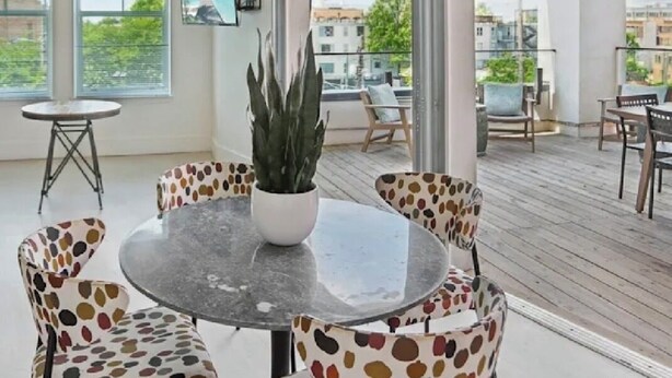 Un espace extérieur aménagé avec une zone de repos couverte, comprenant une table ronde et des chaises, un pot de fleurs et une vue sur des bâtiments et de la verdure.