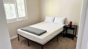 A single bed with a white mattress and pillows, a bedside table with a lamp, and a window with blinds. - Stylish 3BR Kirkland Haven with Backyard Grill (Kirkland)