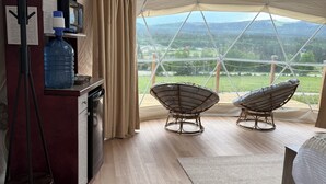 A tented room with a wooden floor, two wicker chairs, a bed, a desk with a water bottle, and a view of a mountainous landscape. - Jasper Gateway Glamping (Hinton)