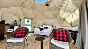 Une chambre aménagée sous une tente, comprenant un lit, deux fauteuils en osier avec des coussins à carreaux rouges et noirs, une petite table et un micro-ondes posé sur une étagère.