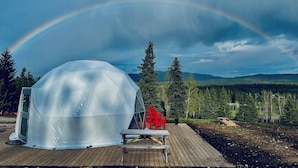 Une structure en forme de dôme transparent, dotée d’une terrasse en bois, d’une chaise rouge et d’un banc.