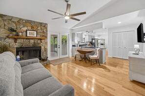 A spacious living area with a stone fireplace, a ceiling fan, a dining table with chairs, and a kitchen in the background. - Quiet Neighborhood, Fenced Yard, Dog Friendly (Fernandina Beach)
